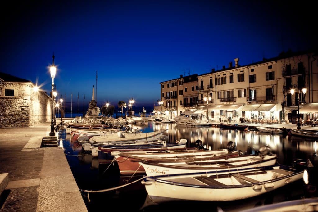 The Venetian harbour of Lazise illuminated at dusk, with boats moored along the quay